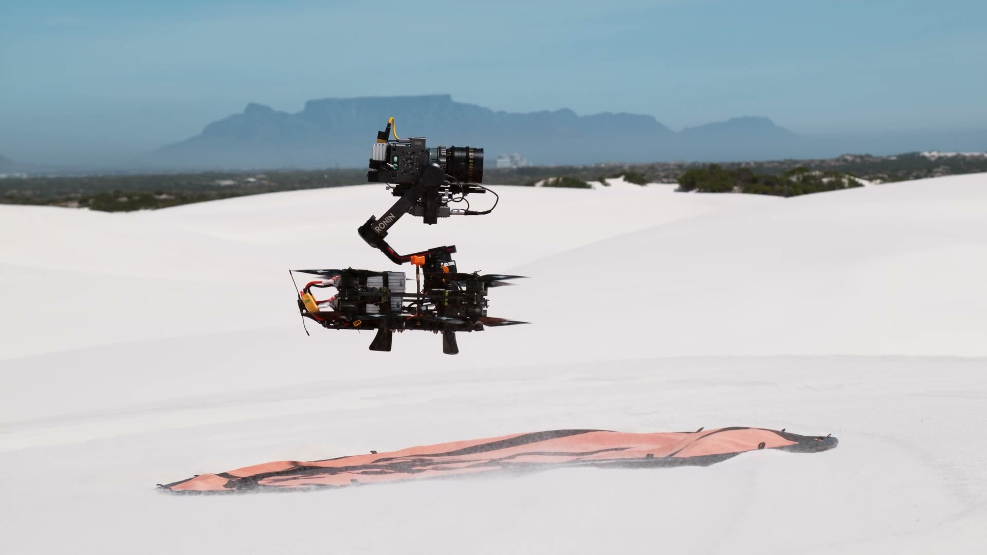 Gimbal-stabilised FPV rig hovering over white dunes, Table Mountain behind